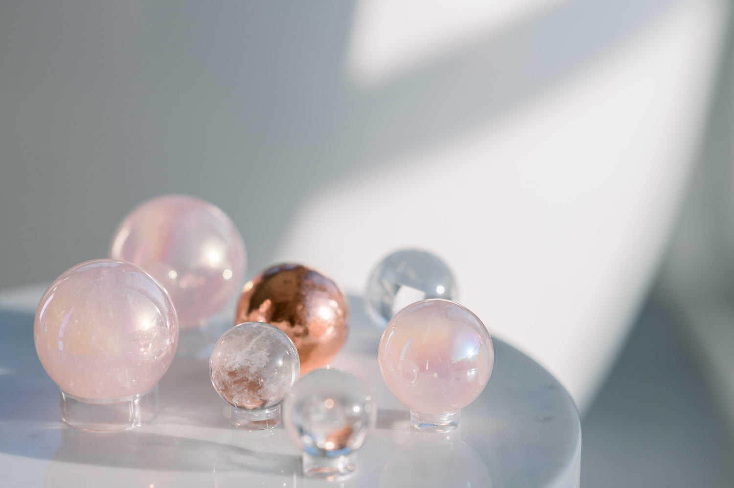 a stone table sits in front of a white background with 7 crystals spheres on top. 3 are small Clear Quartz, one is Copper, and 3 are made of Rose Quartz. All the spheres sit on a clear acrylic stand so they do not roll off the table. 
