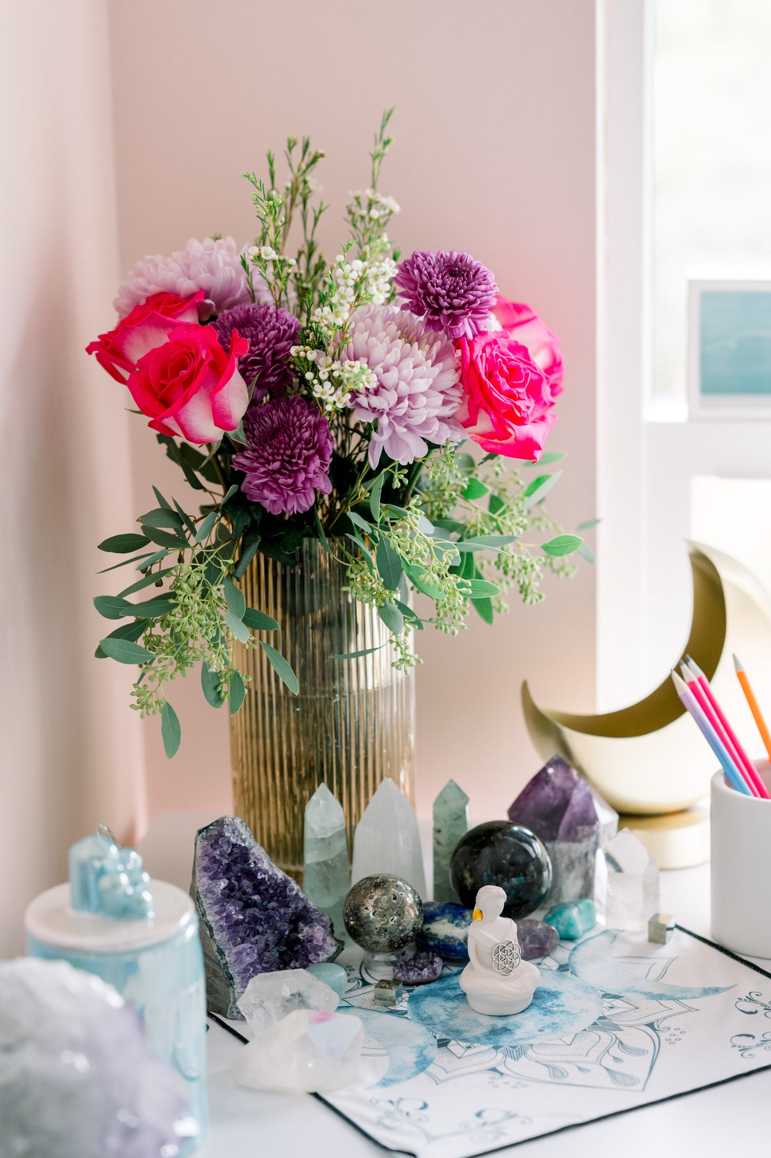assortment of crystals on the desk near a window with a vase of flowers in the background.